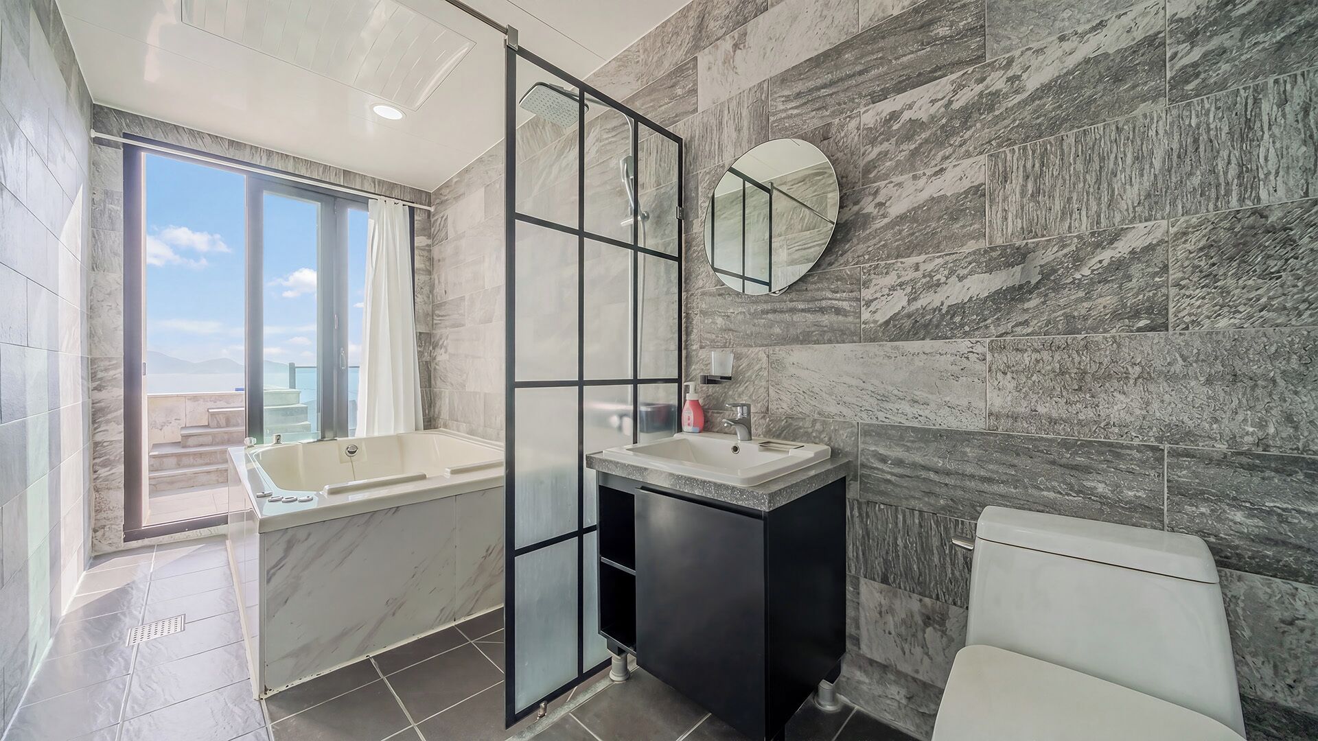 Un baño moderno con bañera, lavamanos e inodoro, con azulejos de mármol y un ventanal grande.