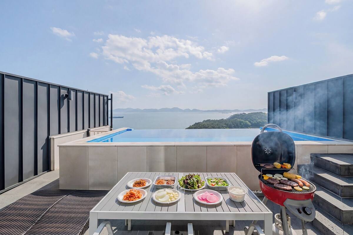Una piscina en la azotea con vistas al mar y a las montañas, una parrilla con comida y una mesa con diferentes platos.