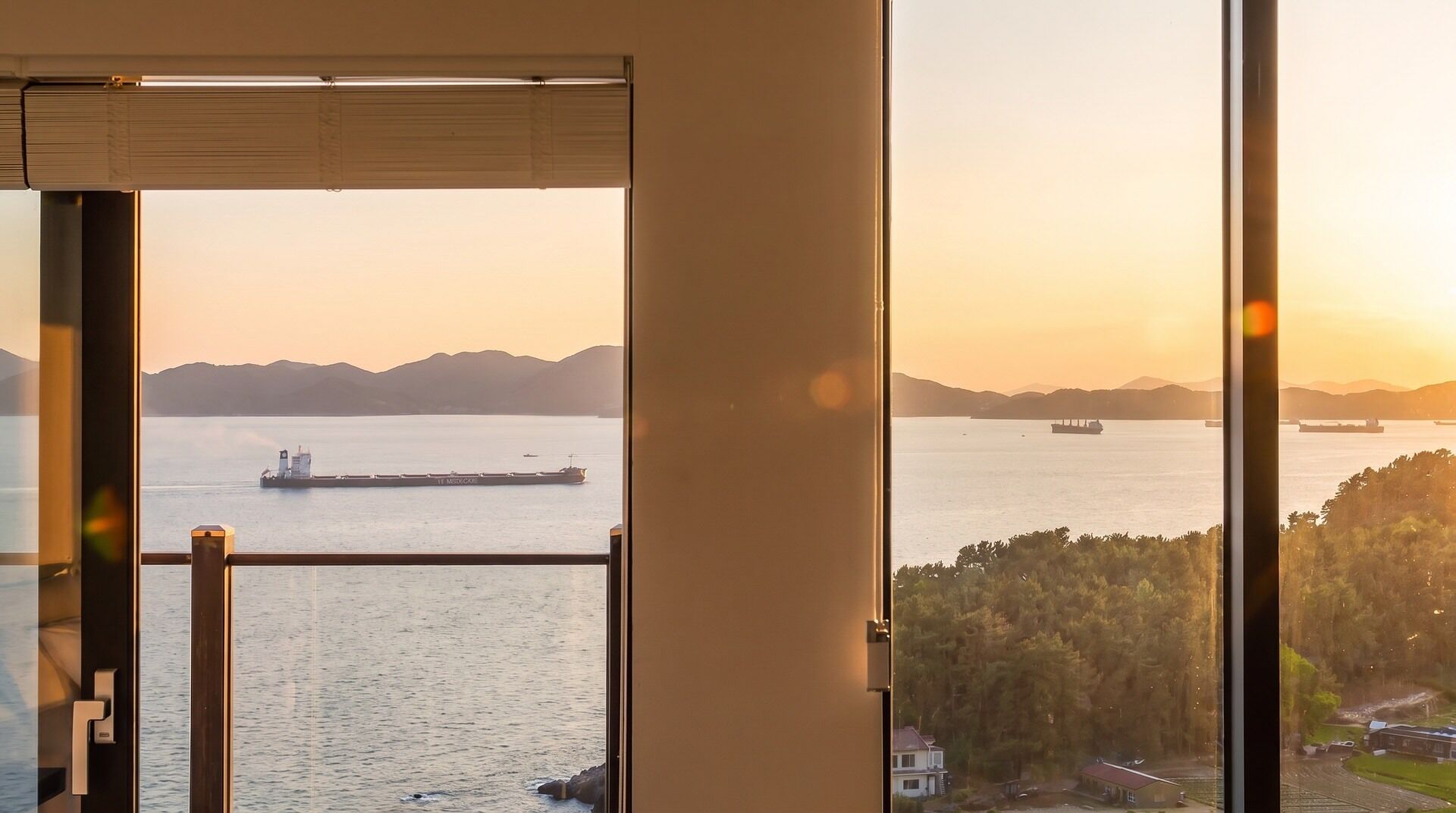 Vista desde una habitación con un gran ventanal que da a una masa de agua con barcos, una zona boscosa y montañas a lo lejos.