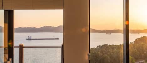 A view from a room with a large window overlooking a body of water with ships, a forested area, and distant mountains.