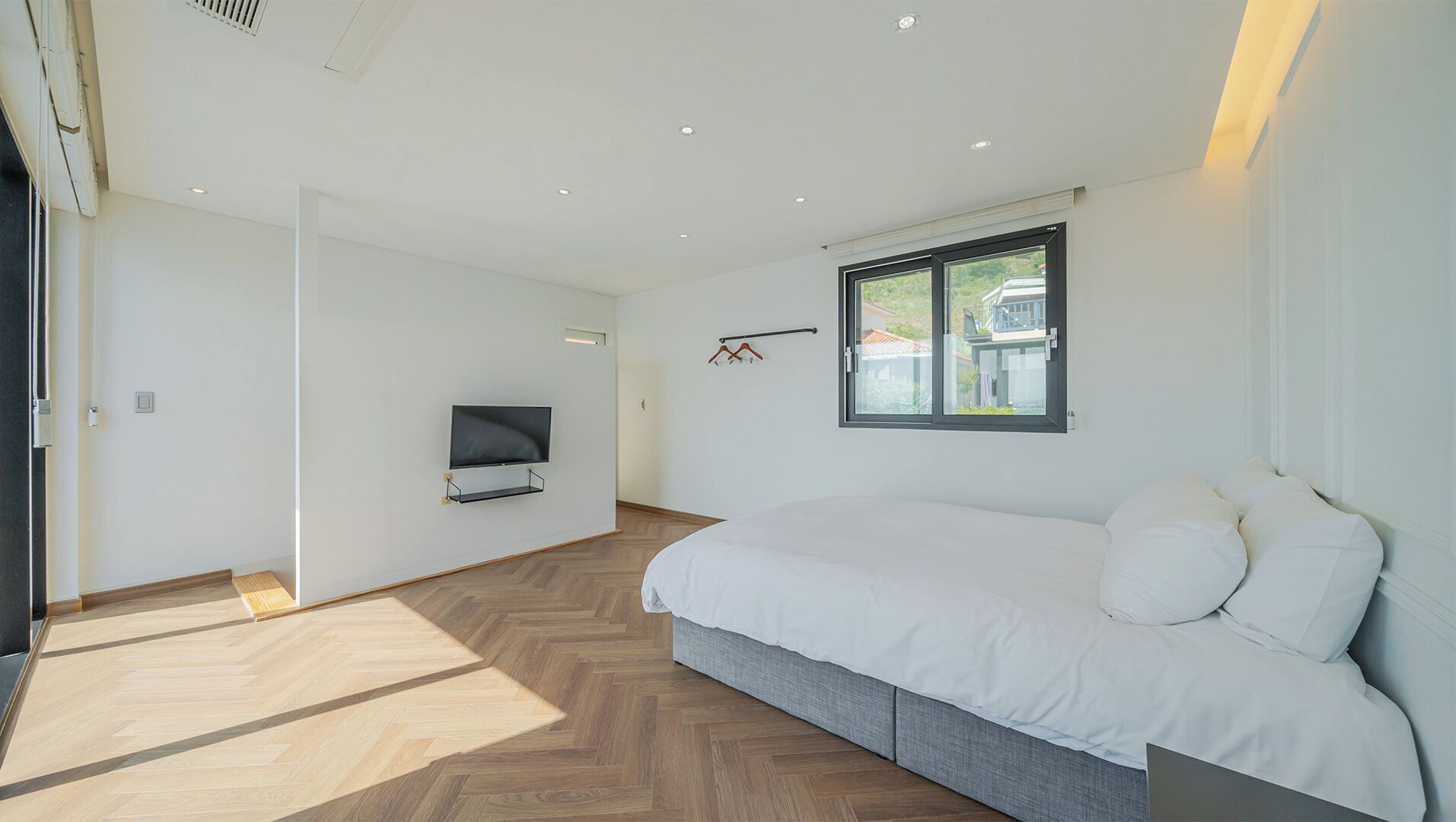 A modern bedroom with a large bed, a TV mounted on the wall, and a window with a view of houses.