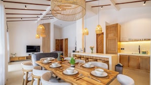 Un comedor moderno con una mesa de madera dispuesta para seis personas, rodeada de sillas blancas, un televisor y una cocina al fondo.