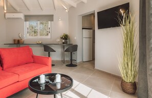 Ein modernes Wohnzimmer mit einem roten Sofa, einem kleinen runden Tisch mit Kerzen, einer Küchenzeile inklusive Kühlschrank und einem an der Wand befestigten Fernseher.