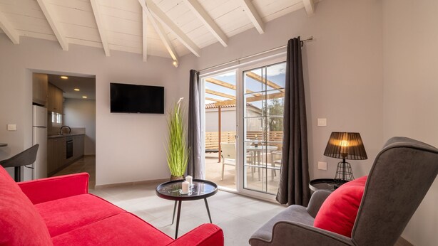 Un salon moderne avec un canapé rouge, un fauteuil gris, une petite table avec une bougie, un téléviseur fixé au mur et une porte-fenêtre coulissante donnant sur un espace extérieur.