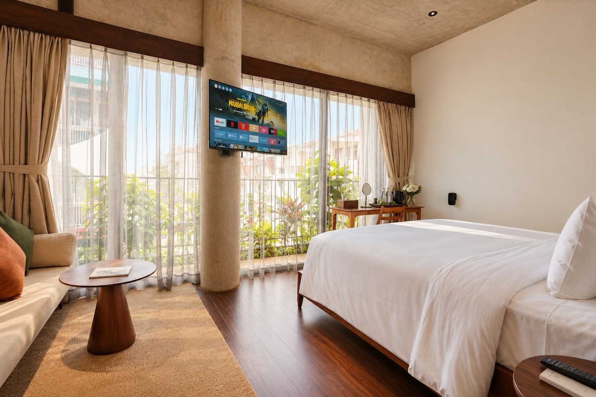 Quarto de hotel com cama, sofá, mesa pequena, TV fixada em suporte e vista para área verde lá fora.
