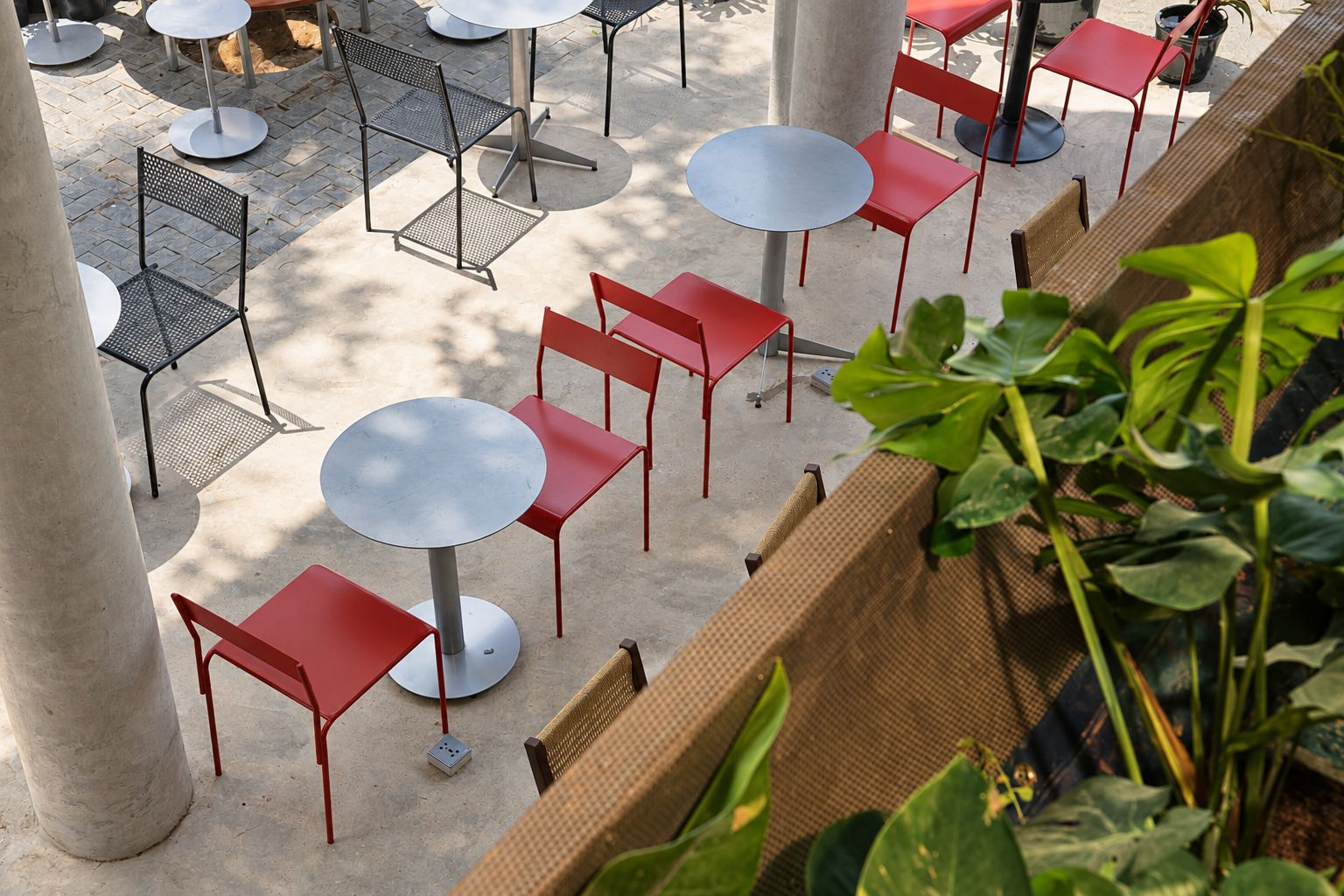 An outdoor seating area with round tables and red chairs, surrounded by concrete pillars.
