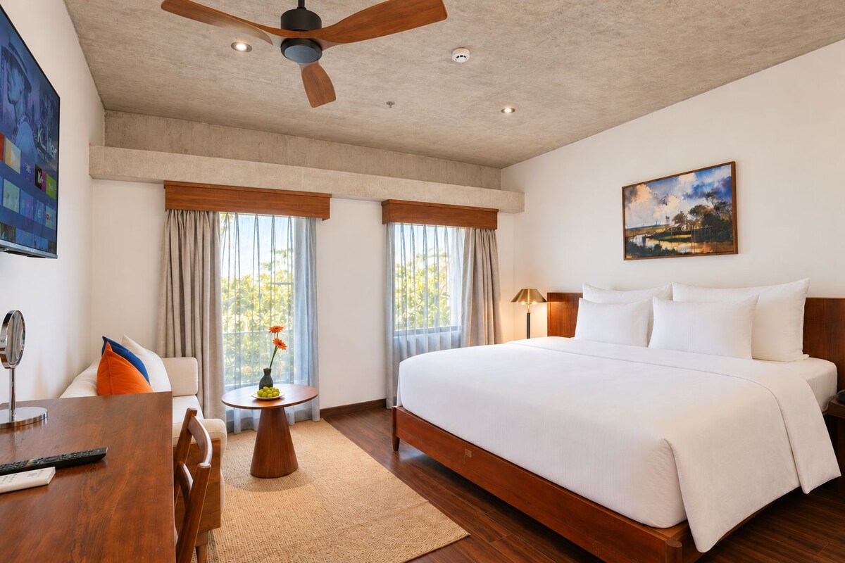 Quarto de hotel com uma cama grande, uma escrivaninha com cadeira, uma mesinha com um vaso e um ventilador de teto.