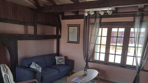 A room with a wooden ceiling, a blue sofa, a white chair, a small table with a lamp, and a window with curtains. - Gite La Granica (restored barn) (Bosset)