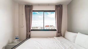 A hotel room with a bed, a window with curtains, a view of buildings, and a water bottle on the nightstand. - Nawin Hotel Donmueang (Bangkok)