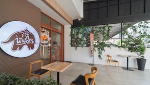 An outdoor seating area with wooden tables and chairs, a large elephant logo, and greenery. - Nawin Hotel Donmueang (Bangkok)