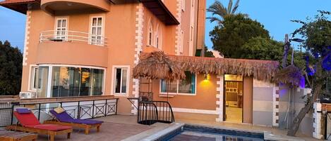 A hotel with a swimming pool, lounge chairs, and a thatched-roof structure.