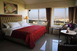 A hotel room with a bed, a chair, a table, a window with a view, and a painting on the wall.