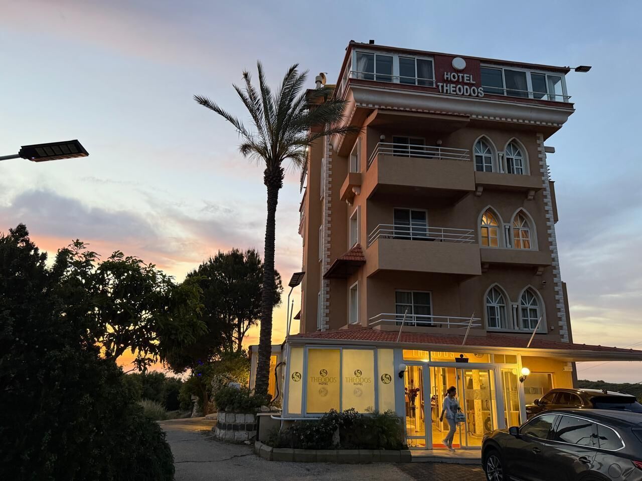 A multi-story hotel with a balcony on the upper floor, a sign reading 'Hotel Theodos,' and a person walking towards the entrance.