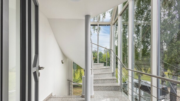 Eine moderne Treppe mit Glasgeländern, die zu einem Außenbereich mit Blick auf Bäume und einen Zug führt.