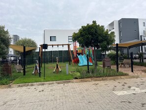 Ein Spielplatz mit Schaukeln und Rutschen, umzäunt, mit Gebäuden im Hintergrund.