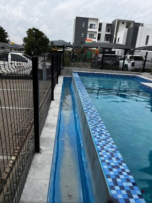 Une piscine dont le bord est recouvert de carreaux, entourée d’une clôture noire, avec des bâtiments en arrière-plan.
