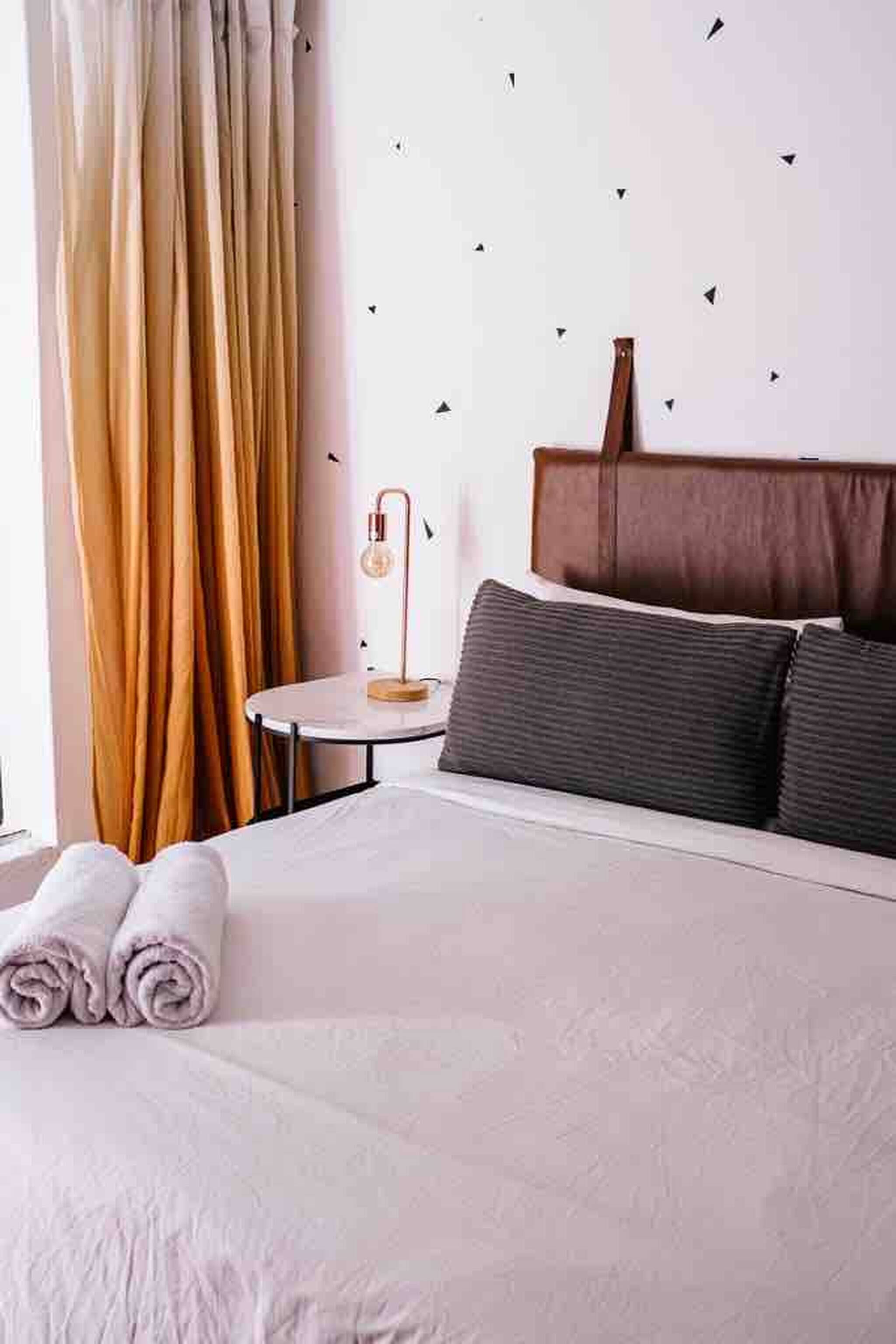 Una cama bien hecha con toallas blancas enrolladas, una mesita redonda con lámpara y un cabecero de madera.