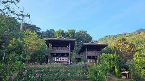 Una estructura de madera tradicional con balcón, rodeada de un jardín con diversas plantas y flores.