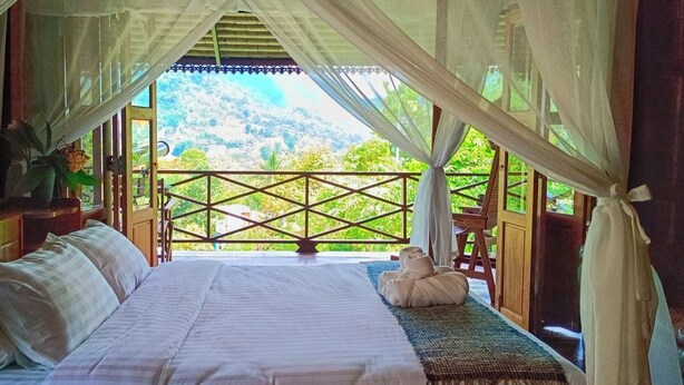 Un dormitorio con cama, almohadas, una mesita de noche, una ventana con vista a la montaña y un balcón con barandilla de madera.