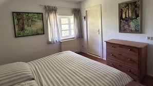 A bedroom with a bed, a dresser, a radiator, a window with curtains, and two paintings on the walls.