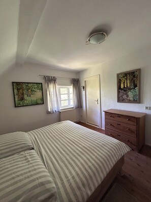 A bedroom with a bed, a dresser, a radiator, a window with curtains, and two paintings on the walls.