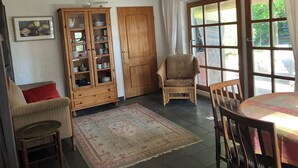 A room with a dining table, chairs, a wicker armchair, a wooden cabinet with glass doors, a patterned rug, and a large window with curtains.