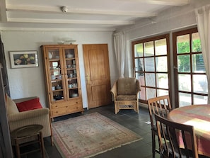 A room with a dining table, chairs, a wicker armchair, a wooden cabinet with glass doors, a patterned rug, and a large window with curtains.
