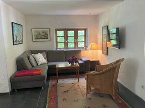 Una sala de estar con un sofá, una silla de mimbre, una mesa con un jarrón, una lámpara y una ventana con vista a zonas verdes.