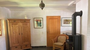 A room with a wooden wardrobe, a wicker chair, a black stove, and two framed pictures on the wall.