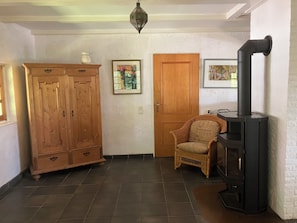 A room with a wooden wardrobe, a wicker chair, a black stove, and two framed pictures on the wall.