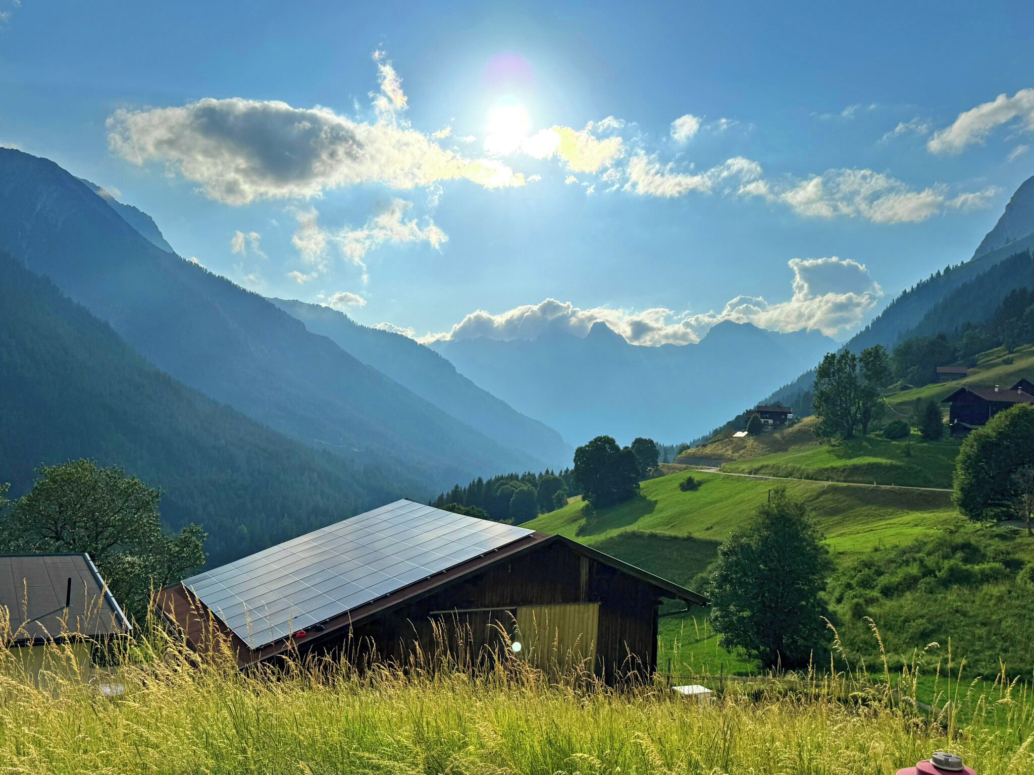 Ländliche Landschaft mit Bergen, einem Holzhaus mit Solardach und blauem Himmel.