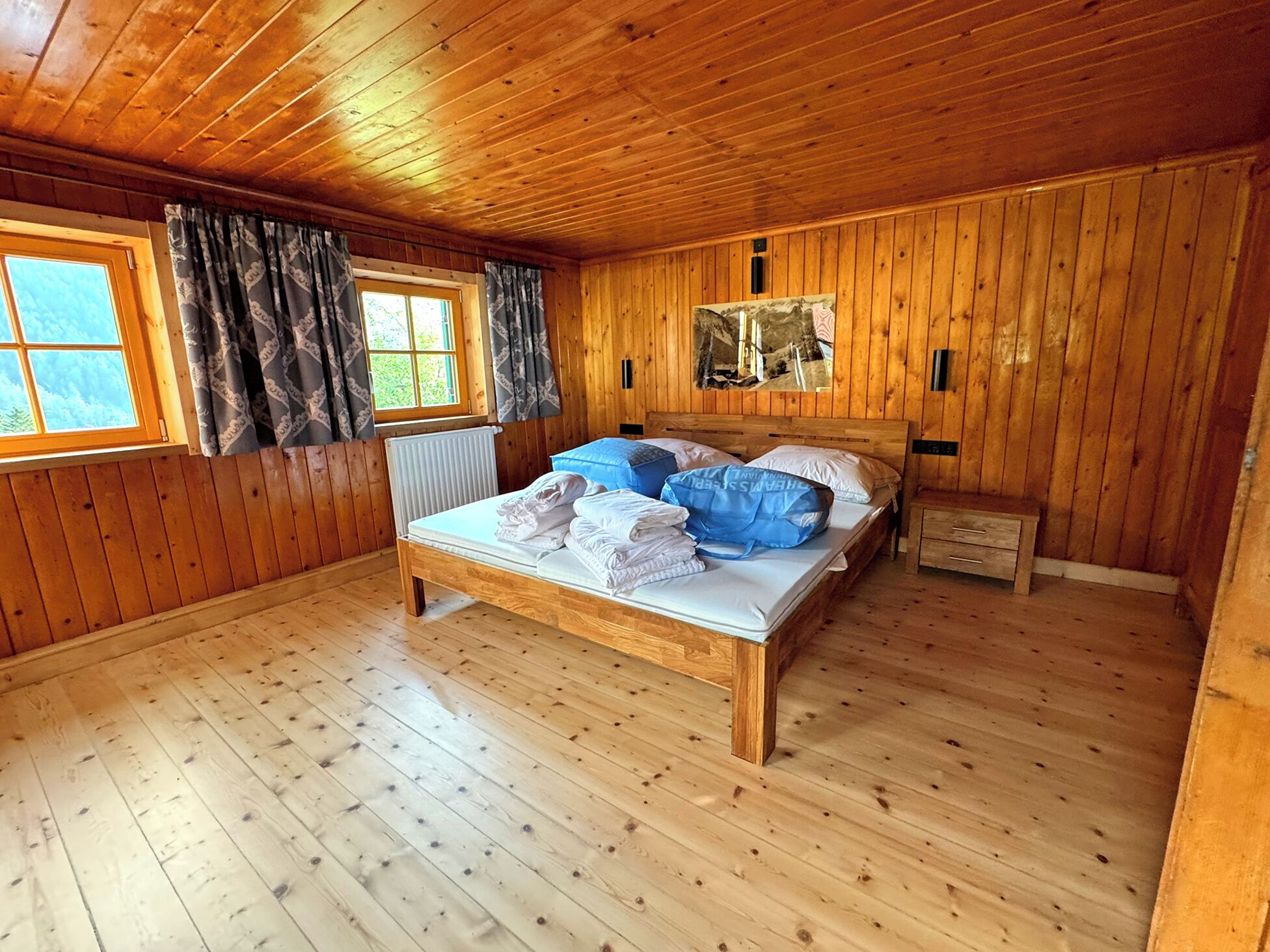 Una habitación con paneles de madera, una cama, una ventana con cortinas, un radiador y una mesita de noche pequeña.
