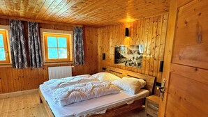 A wooden cabin bedroom with a bed, two windows with curtains, a radiator, and a wall-mounted light fixture.