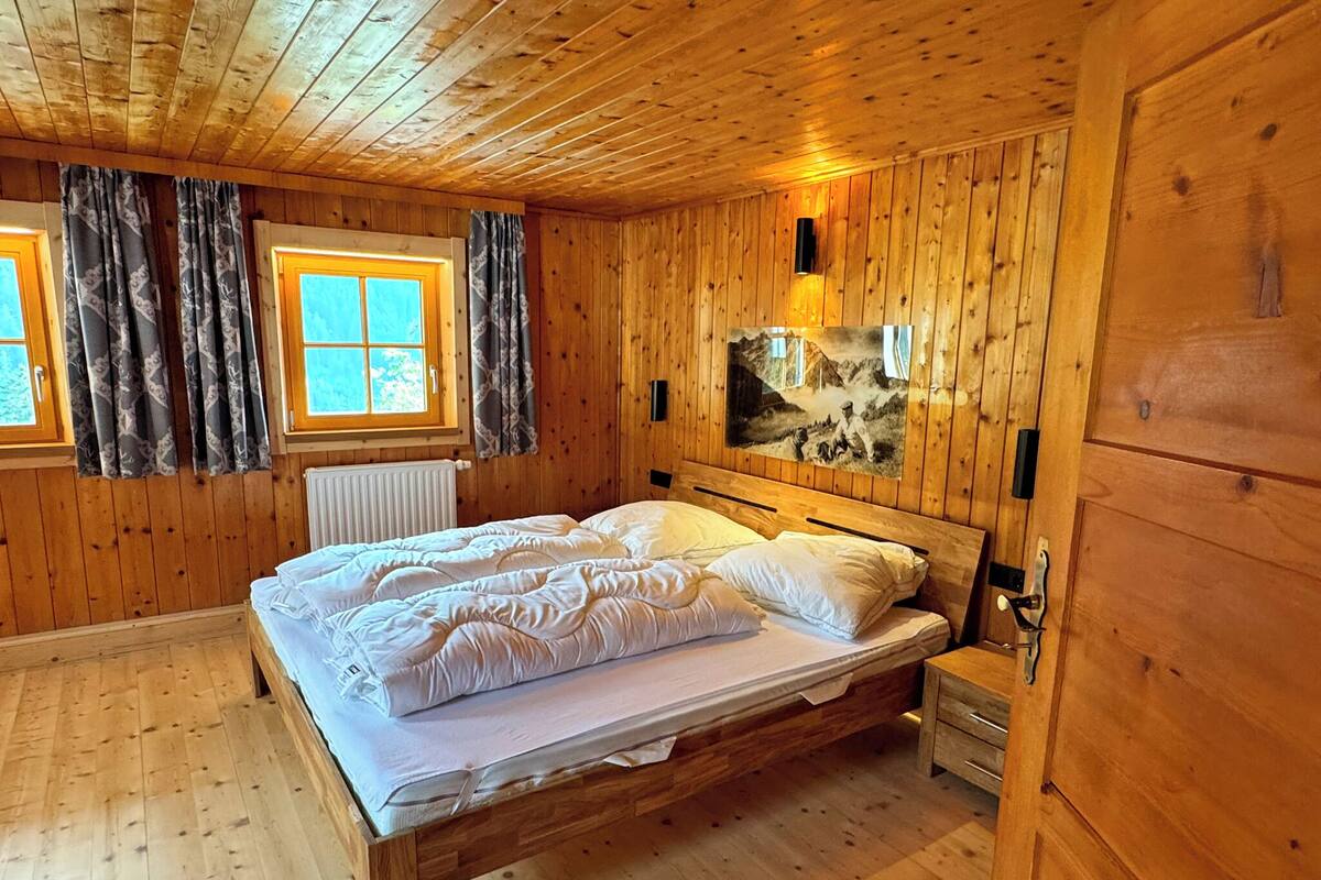 A wooden cabin bedroom with a bed, two windows with curtains, a radiator, and a wall-mounted light fixture.
