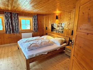 A wooden cabin bedroom with a bed, two windows with curtains, a radiator, and a wall-mounted light fixture. - Zum Hubertus (Pfafflar)