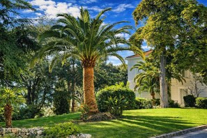 A well-maintained garden with palm trees, a stone wall, and a building with a white facade. - Superb renovated T2 - air conditioning - terrace - swimming pool - tennis court (Cannes)