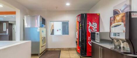 A kitchen area with a refrigerator, a microwave, a Coca-Cola vending machine, and a coffee machine.