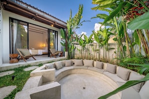 A curved outdoor seating area with a concrete fire pit, surrounded by lush tropical plants and a house with large windows. - Berapi Villa Ubud  (Ubud)