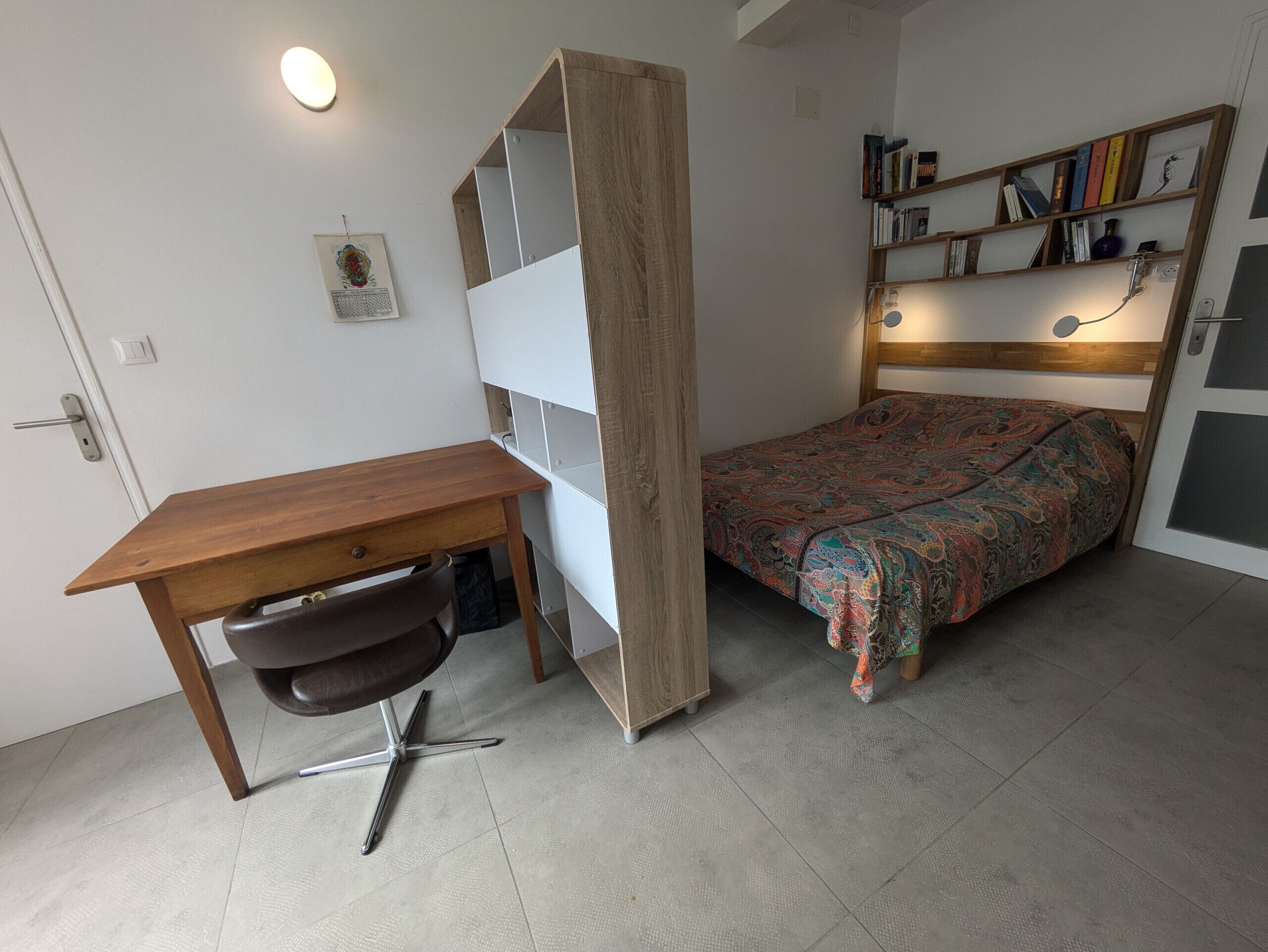 Une chambre à coucher de taille modeste, avec un bureau en bois, un lit recouvert d’une couvre-lit à motifs, une bibliothèque et une petite étagère fixée au mur.