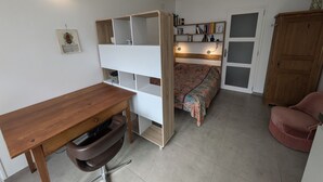 Quarto com escrivaninha de madeira, uma cadeira, cama com colcha estampada, estante de livros e um armário.