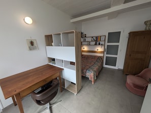 Ein Zimmer mit einem Holztisch, einem Stuhl, einem Bett mit gemustertem Bettbezug, einem Bücherregal und einem Schrank.