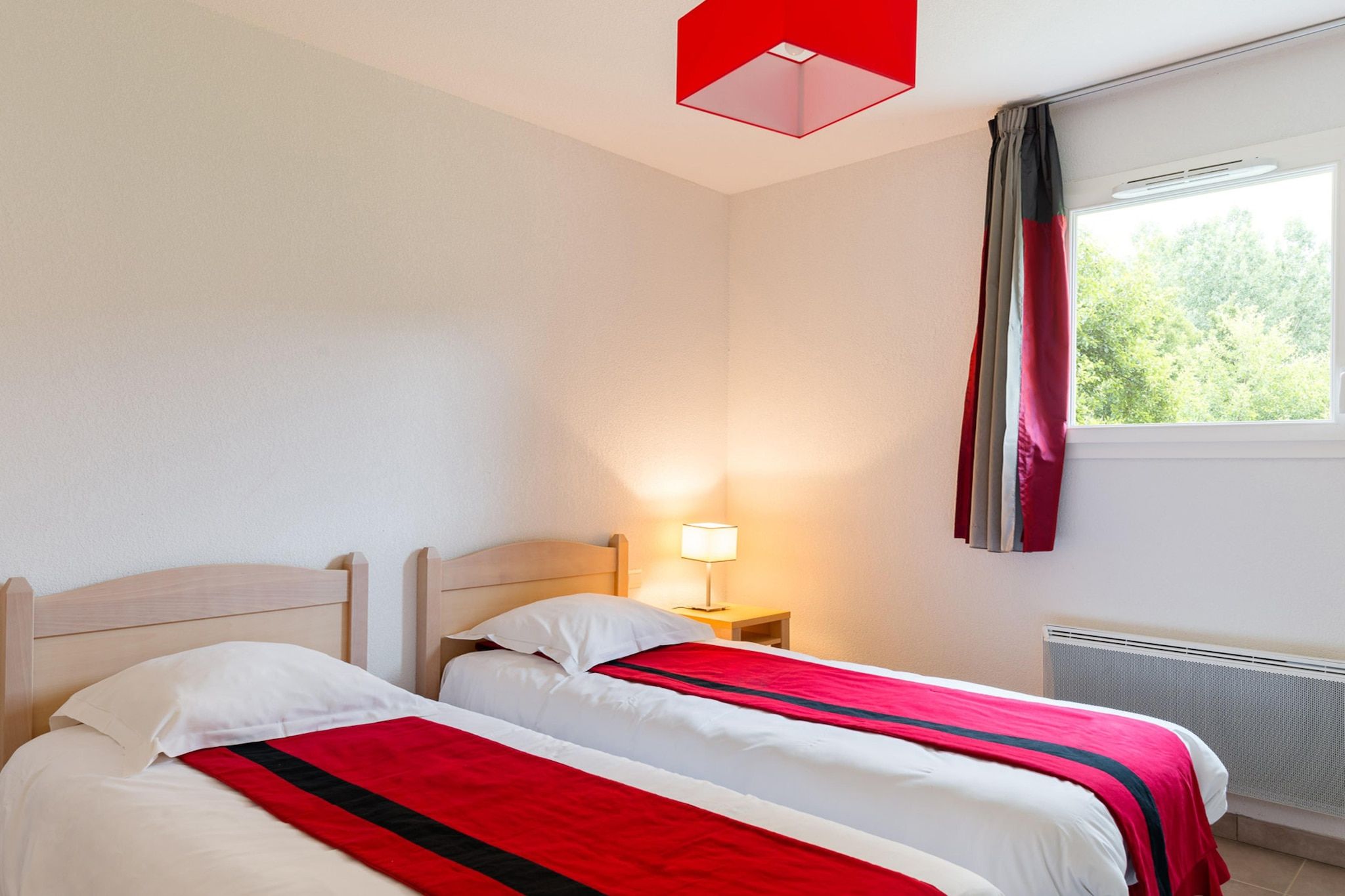 Une chambre d’hôtel avec deux lits, une table de chevet avec une lampe, une fenêtre avec des rideaux et un radiateur.