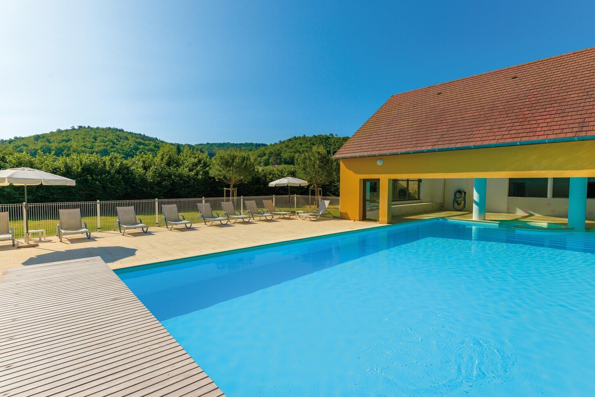 Una piscina con lettini e ombrelloni, un edificio giallo con tetto rosso e una collina ricoperta di alberi sullo sfondo.