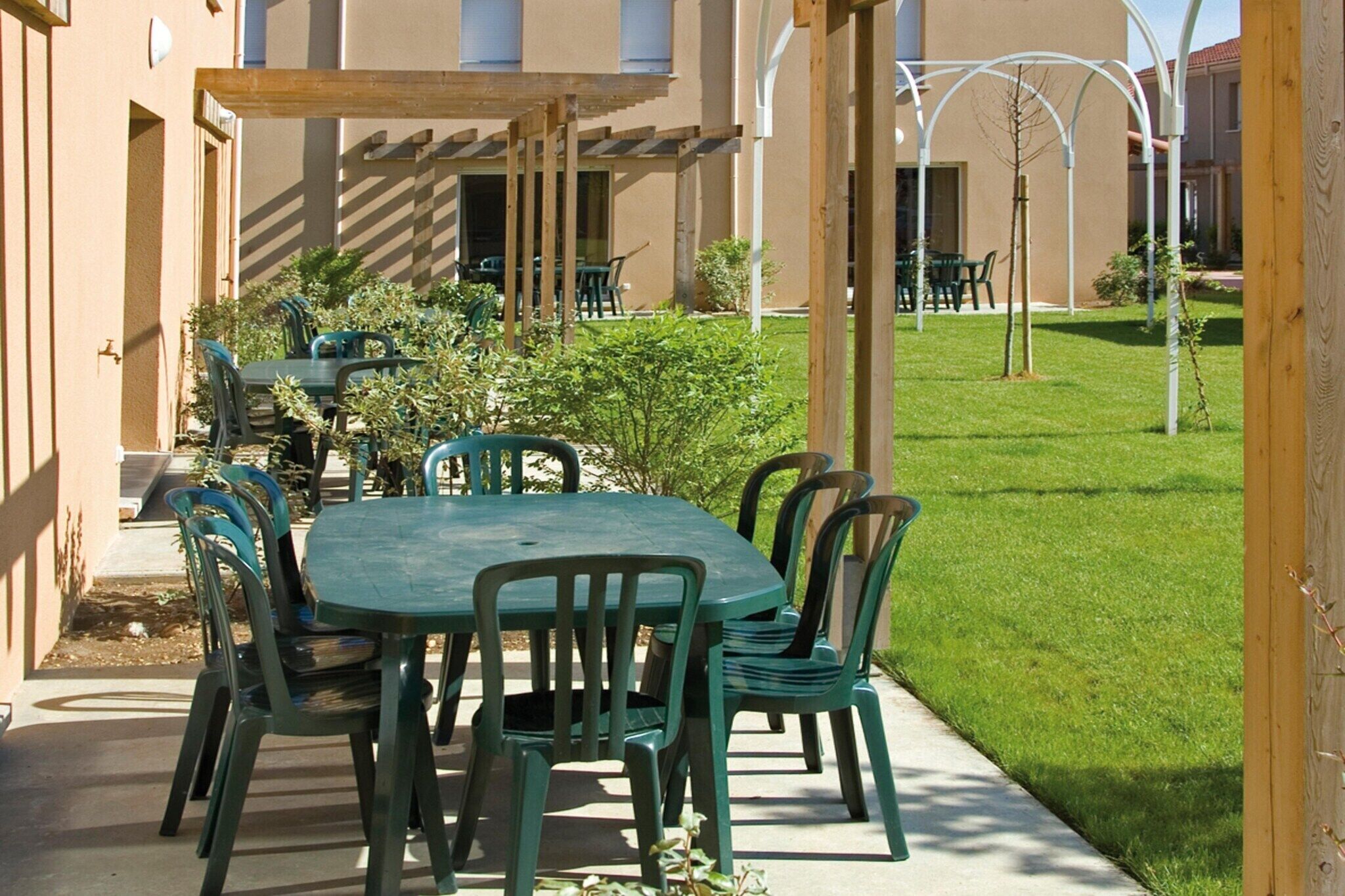 An outdoor patio with a table and chairs, surrounded by greenery and a building in the background.