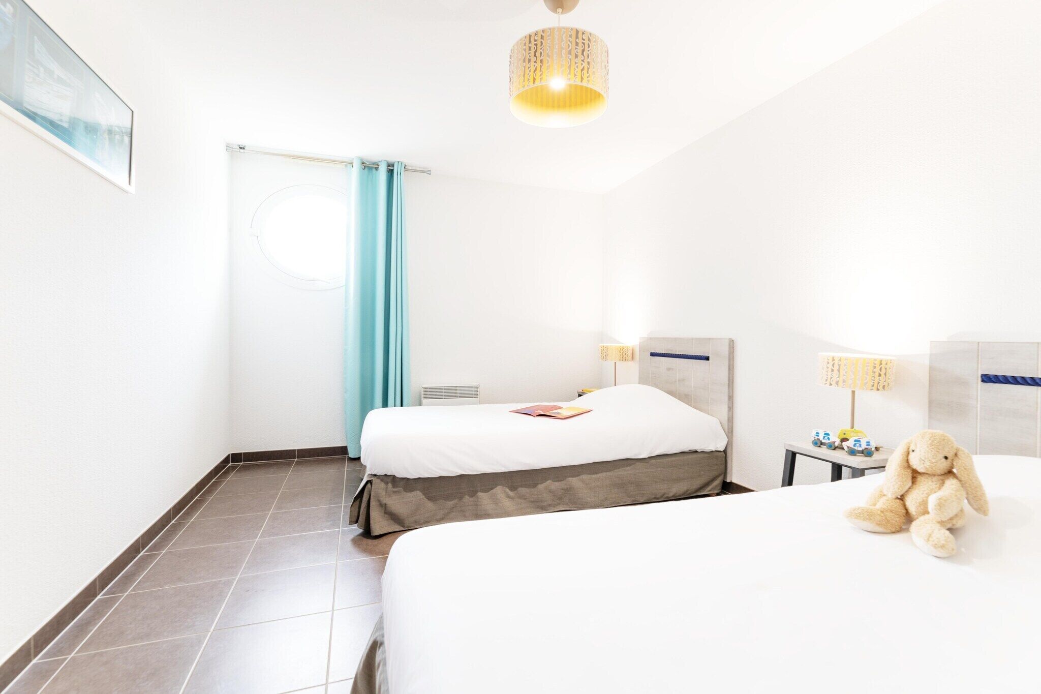 Une chambre d’hôtel avec deux lits, une fenêtre avec des rideaux, une lampe et un ours en peluche.