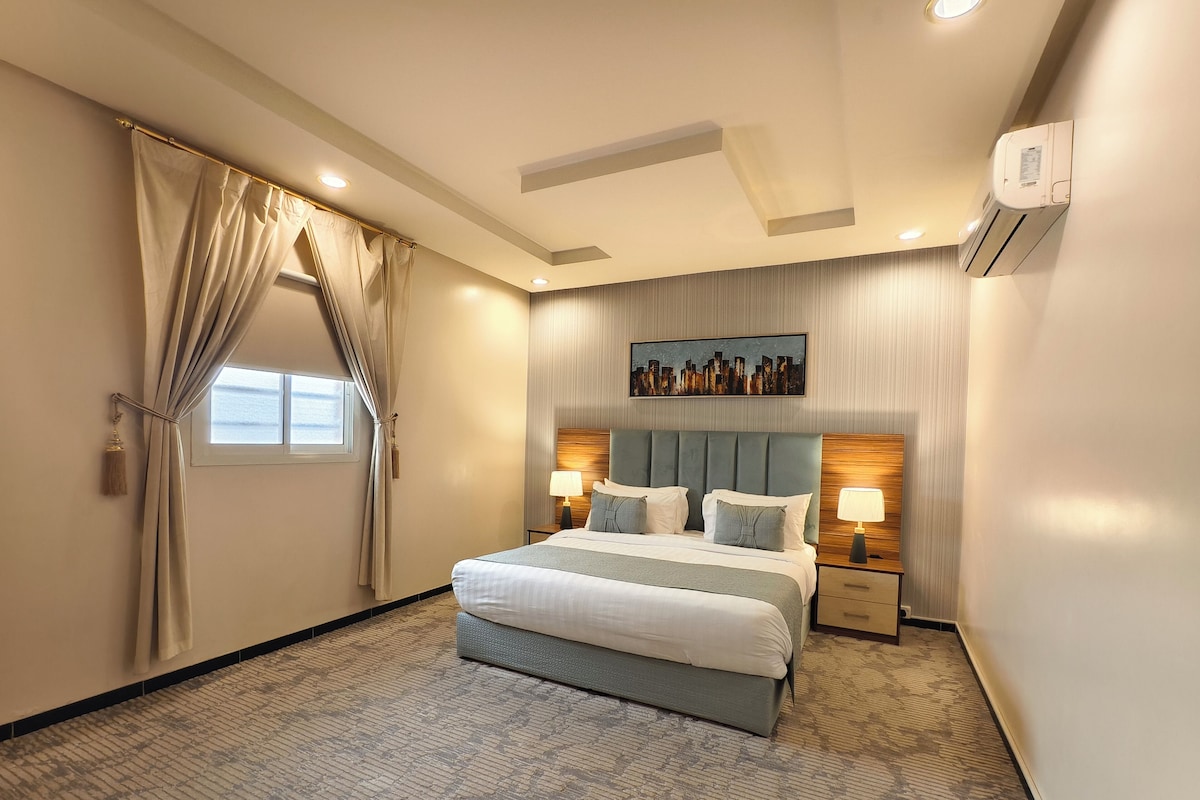 Quarto de hotel com uma cama grande, duas mesas de cabeceira com luminárias, ar-condicionado no teto e uma janela com cortinas.