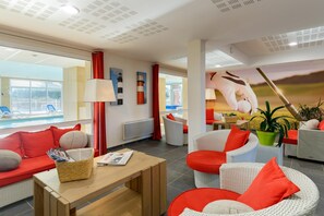 A hotel lobby with a seating area, a table with a magazine, a lamp, and a view of the pool. - Stylish coastal apartment with shared pool (Fréhel)