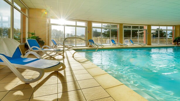 Ein heller, überdachter Pool mit klarem, blauem Wasser, umgeben von sonnendurchfluteten Fliesen und blauen Liegestühlen.