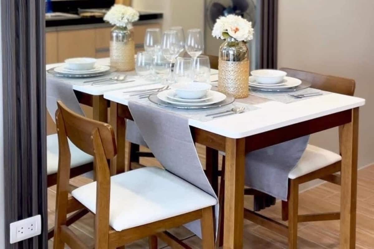 Comedor con mesa dispuesta para cuatro personas, sillas de madera, lámparas colgantes y un florero con flores blancas.