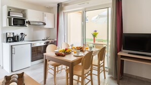 Cocina compacta con microondas, horno y cafetera. Una mesa de comedor con sillas dispuesta para el desayuno. Un televisor sobre un mueble a la derecha.